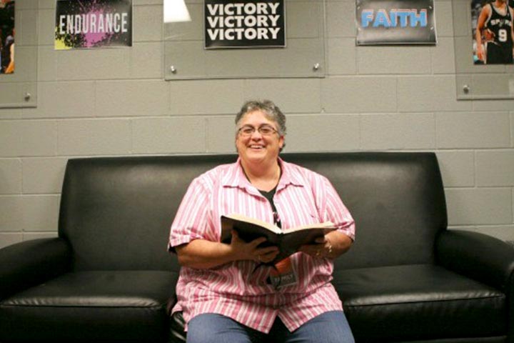 Betsy Buhler Chaplain sitting down on a couch smiling and holding book in front of basketball pictures